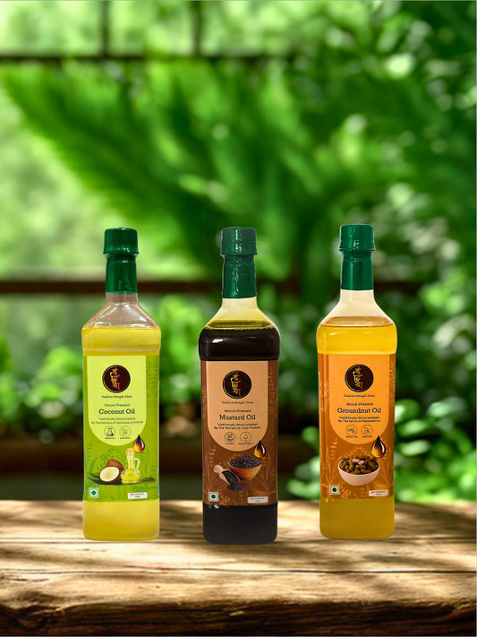 Three bottles of coconut, mustard, and groundnut oils arranged on a rustic wooden table with a blurred green garden background.