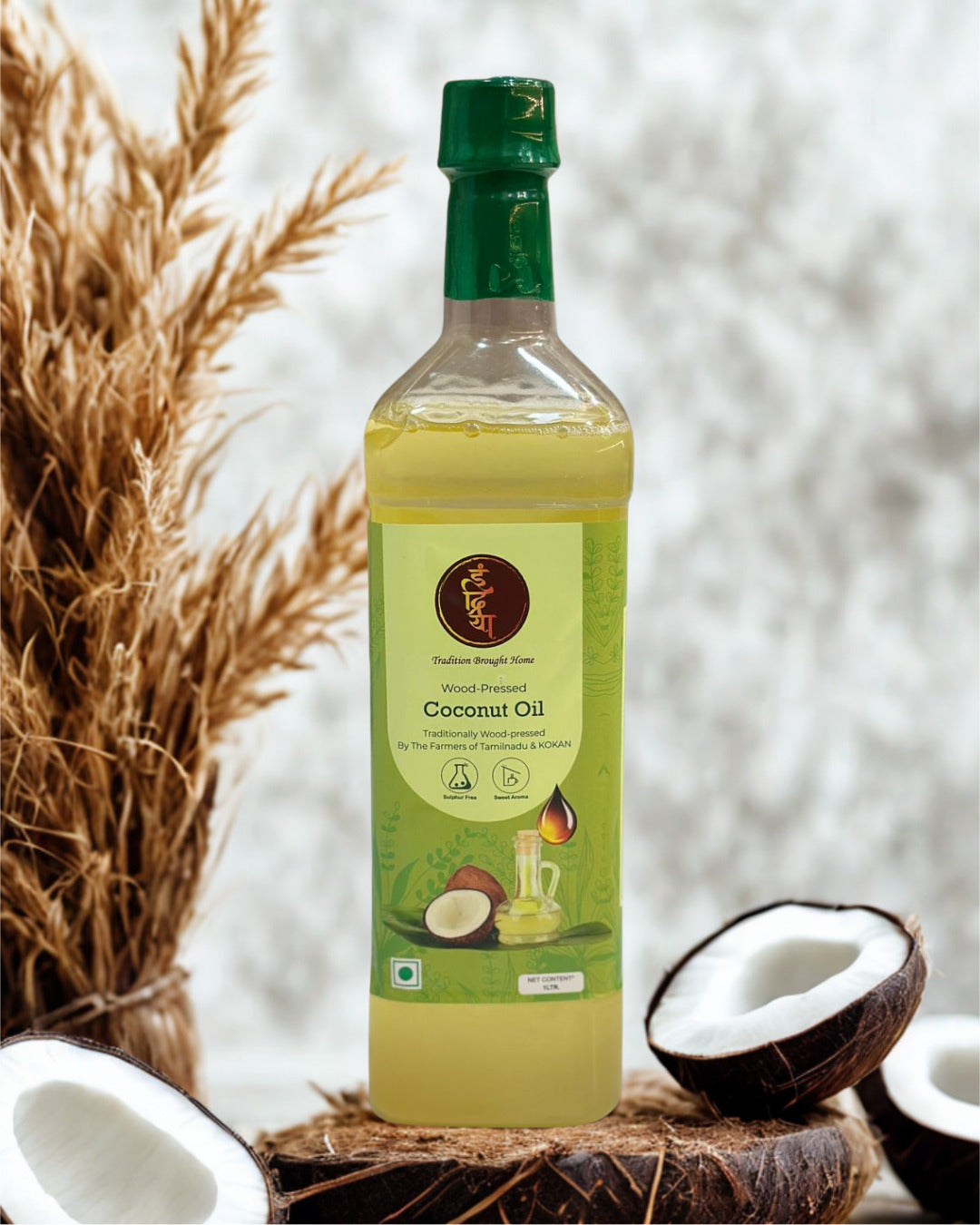 A bottle of Southern Banglaa Coconut Oil with a green cap and green label, styled on a coconut stump with dried grass and halved coconuts against a soft white background.